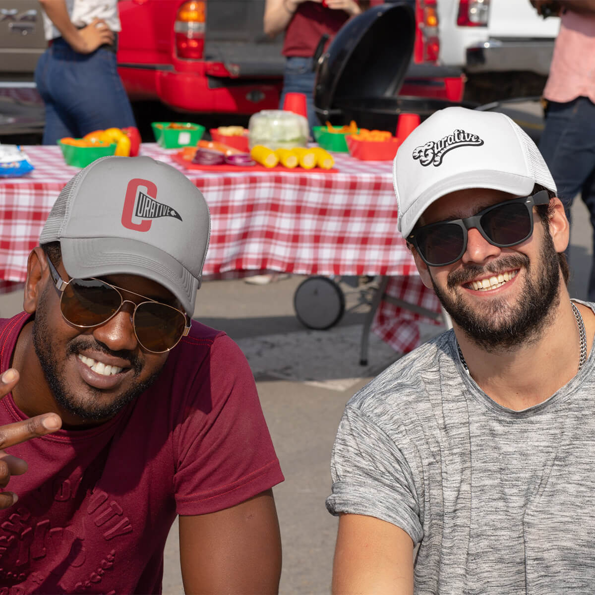 Two men at a tailgate party wearing grey and white trucker hats with black curative printing logo imprints