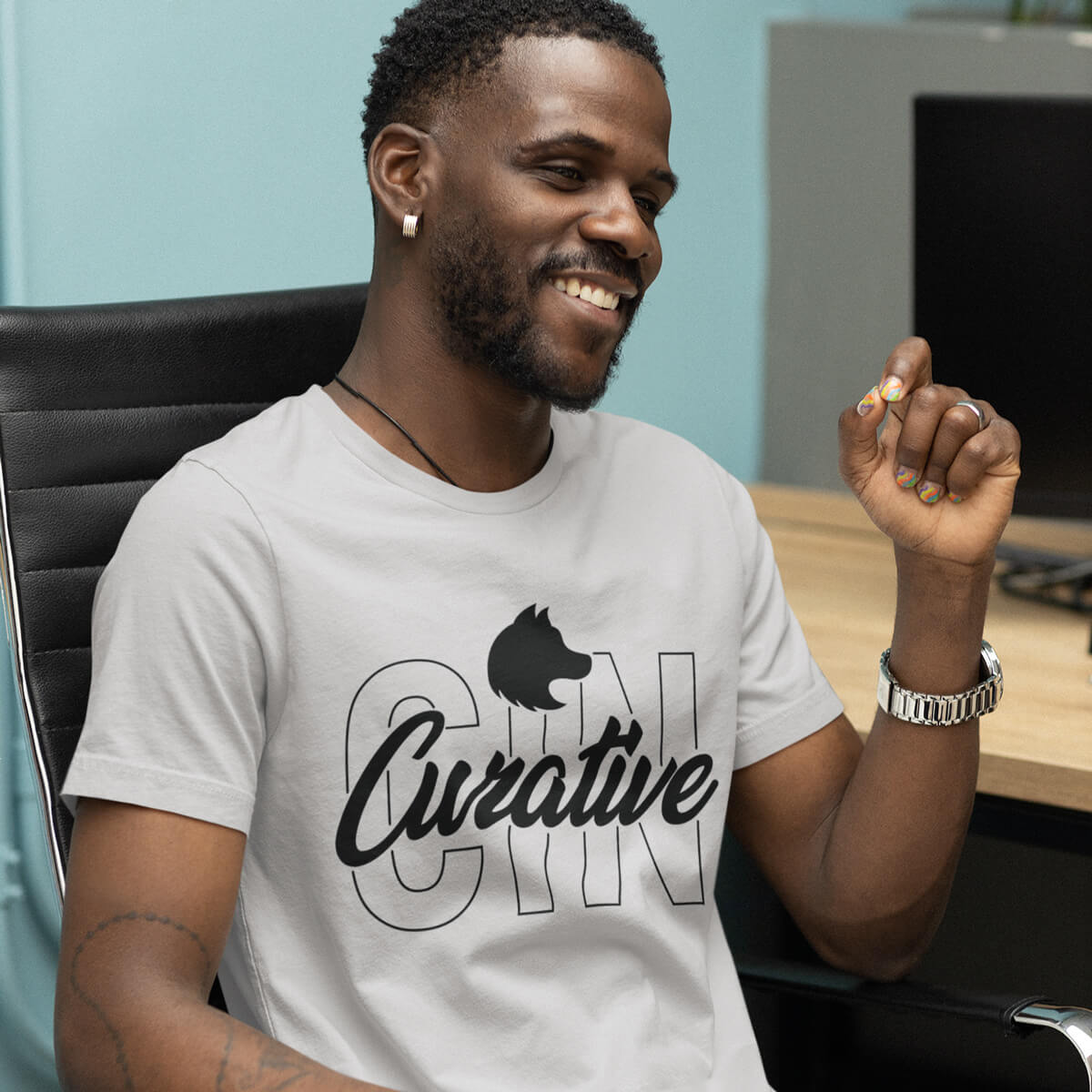 Man sits in desk chair wearing a white short sleeve t-shirt tee with black curative printing logo imprint