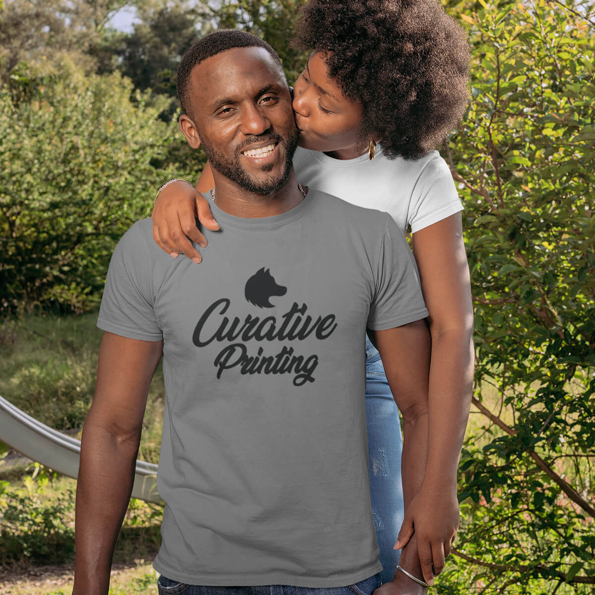 Woman stands behind man and kisses him on the cheek as he wears grey short sleeve t-shirt tee with black curative printing logo imprint