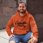 Man crouching against wall wearing an orange pullover hoodie sweatshirt with black curative printing logo imprint