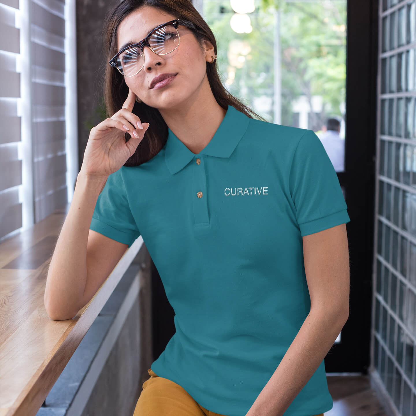Woman sits in a cafe wearing a teal polo collar shirt with white curative printing logo imprint