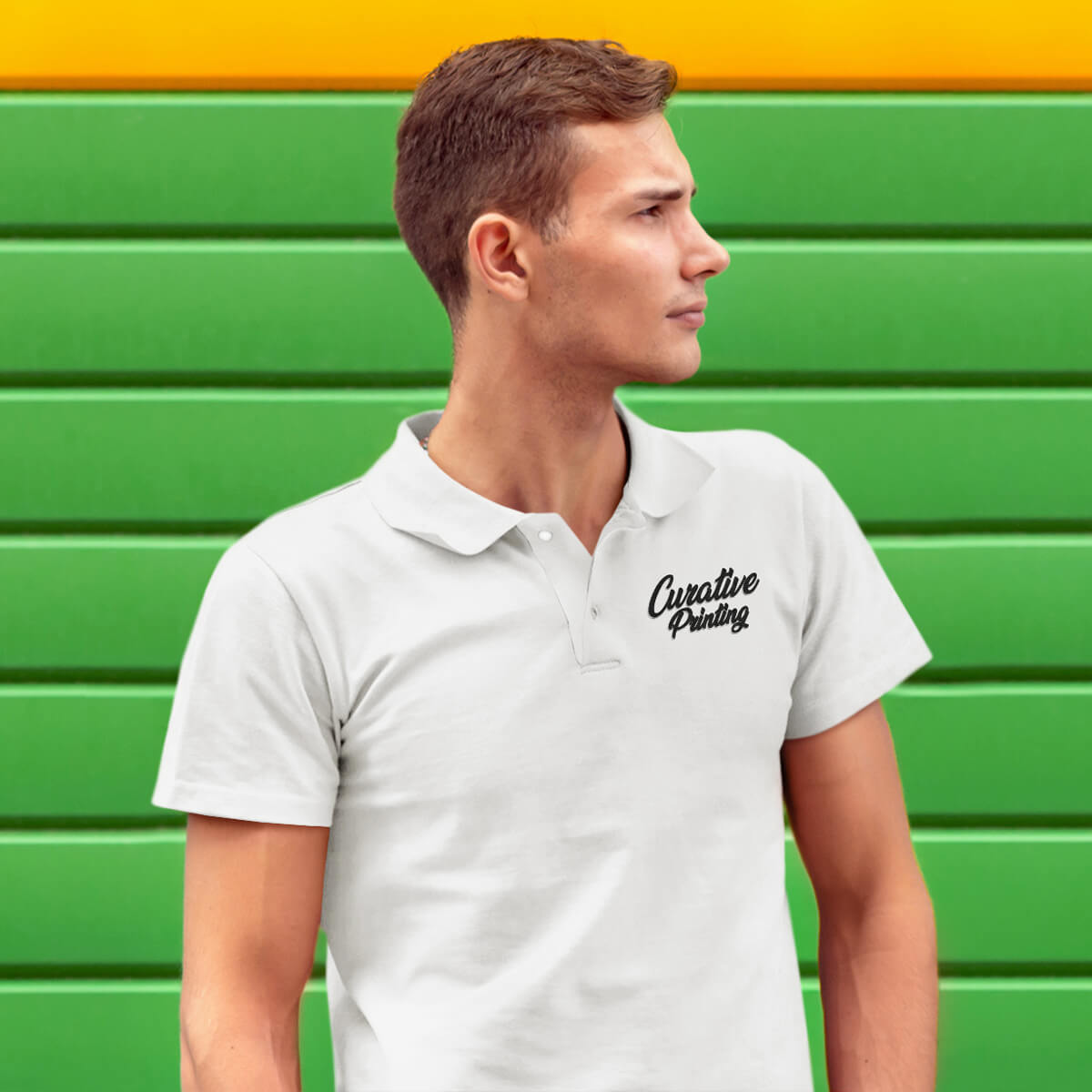 Man stands outside against a green wall wearing a white polo collar shirt with black curative printing logo imprint