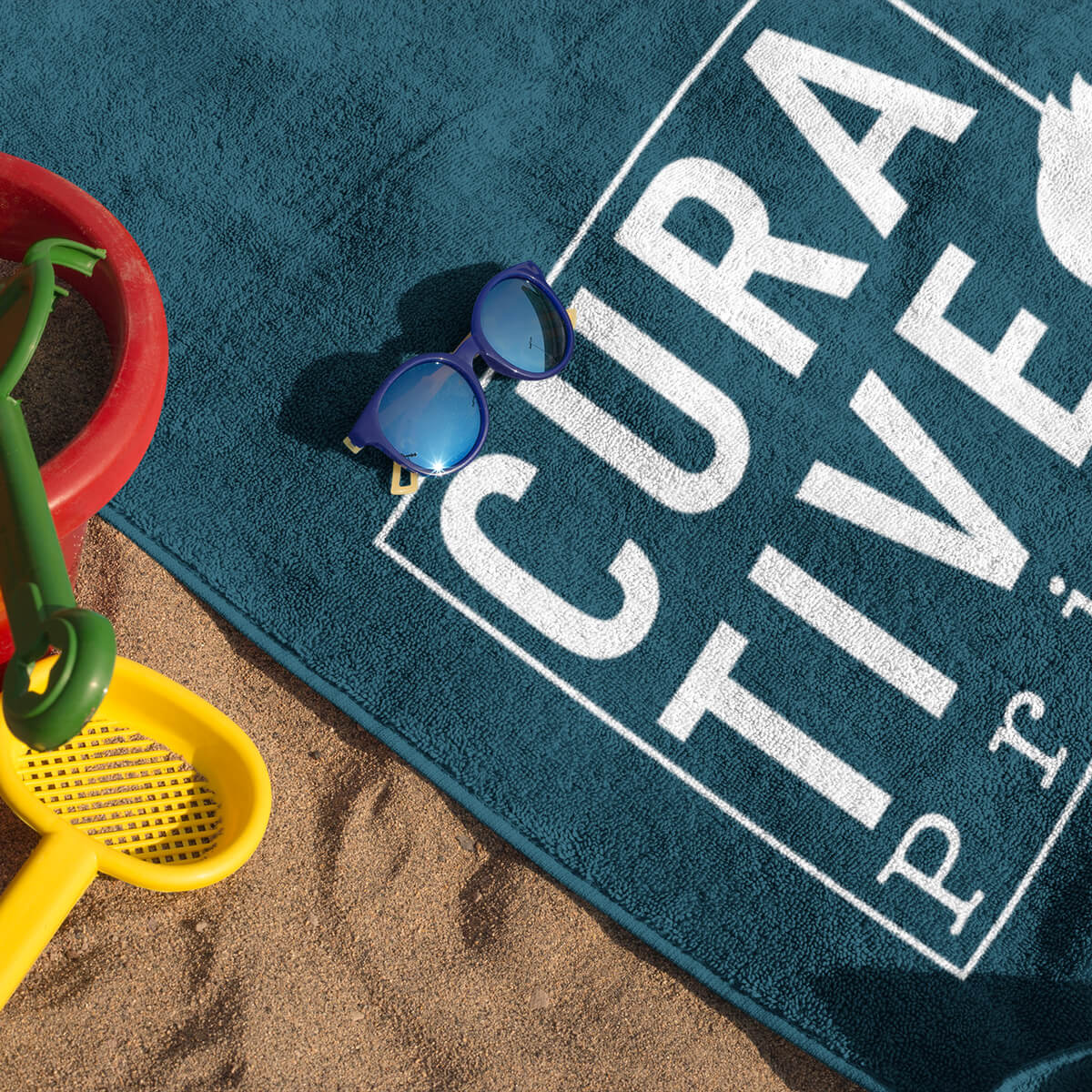 Sand toys, sunglasses next to navy with white imprint beach towel promotional towels by curative printing