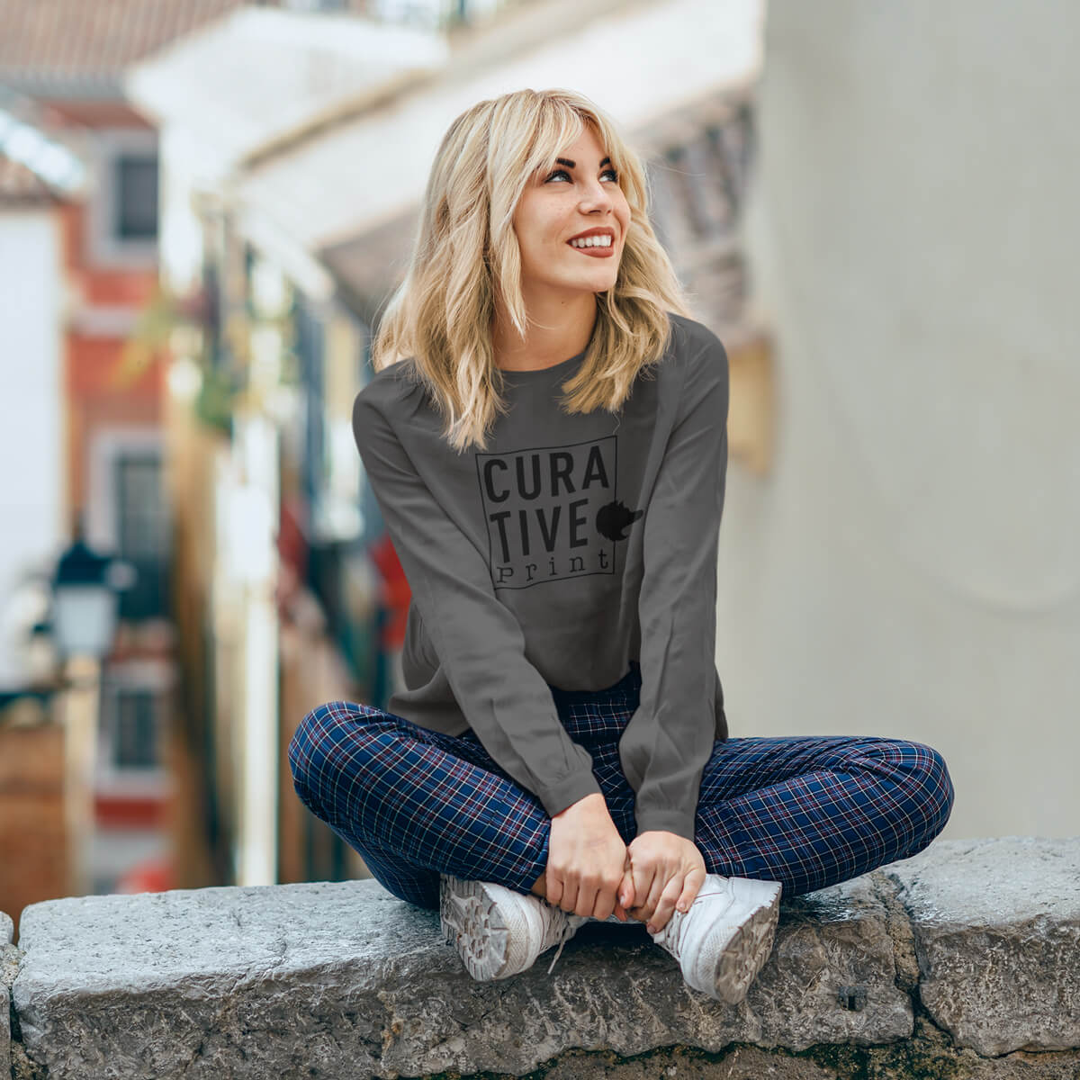 Lady sits on a stone wall wearing a grey long sleeve t-shirt tee with black curative printing logo imprint