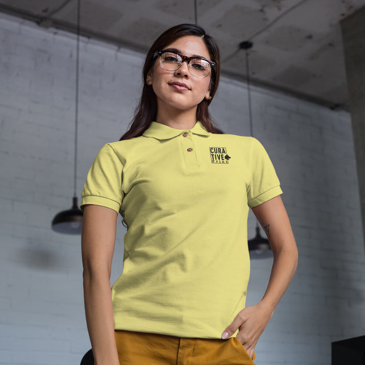 Woman stands in an industrial space wearing a yellow polo collar shirt with black curative printing logo imprint